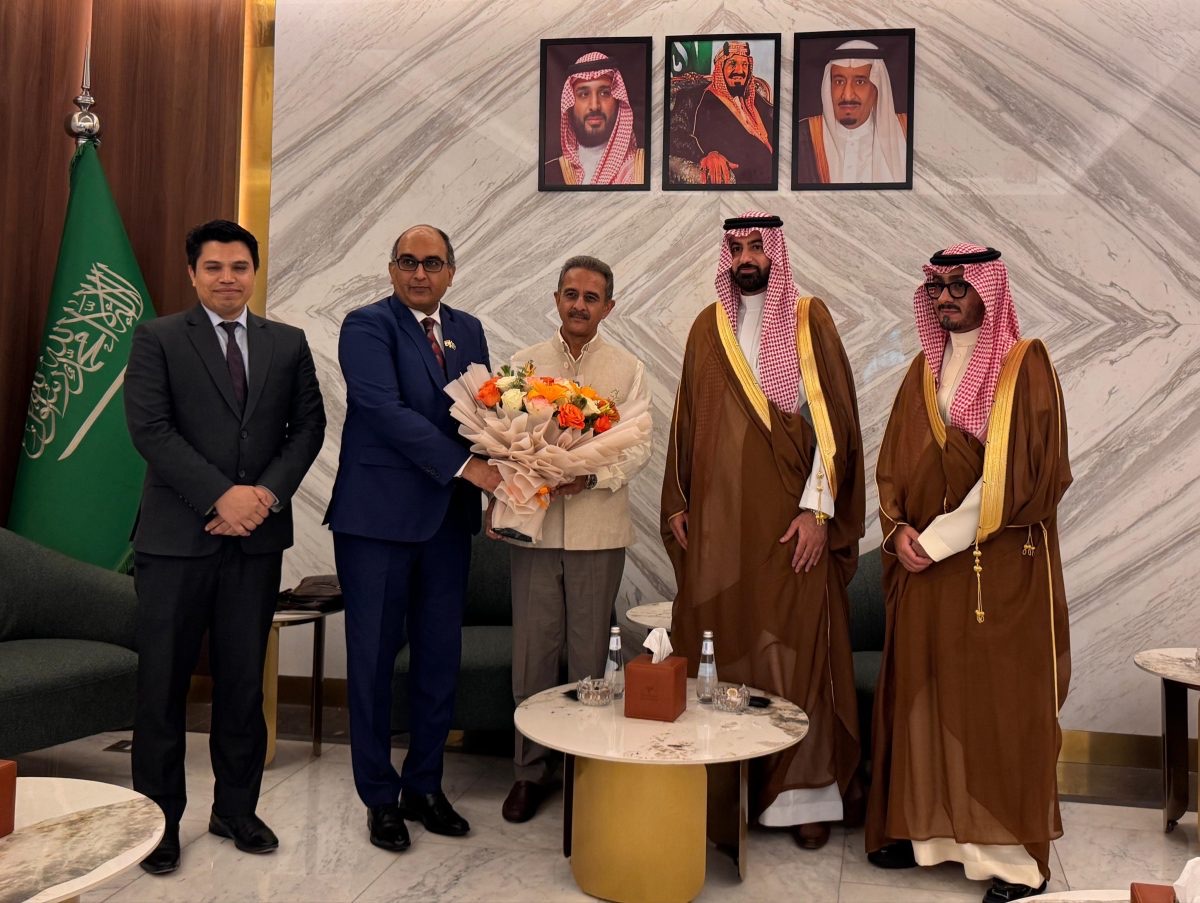 Kirtivardhan Singh holding a bouquet, standing with Indian and Saudi officials beneath portraits of the Saudi royal family and beside the flag of Saudi Arabia
